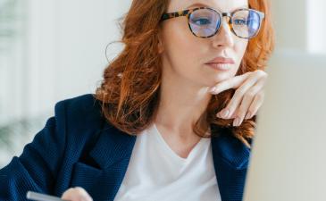 Young woman with red hair and glasses looking at her laptop, thinking while writing in a notebook